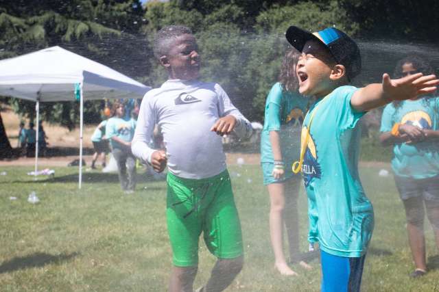 Children playing outdoors in a park, enjoying water spray during a summer activity with others in the background.