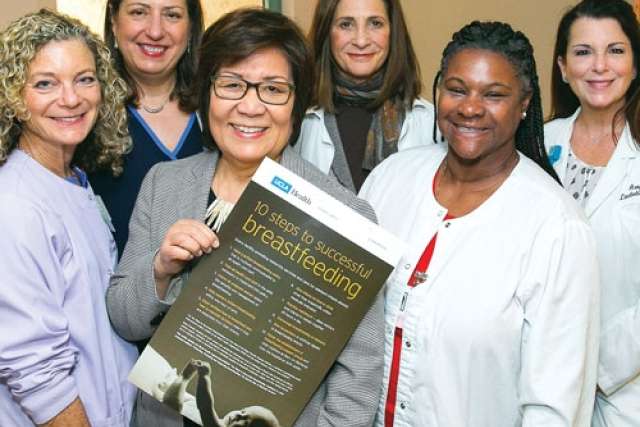 Group of people standing together indoors, holding a large informational poster titled ‘10 steps to successful breastfeeding.