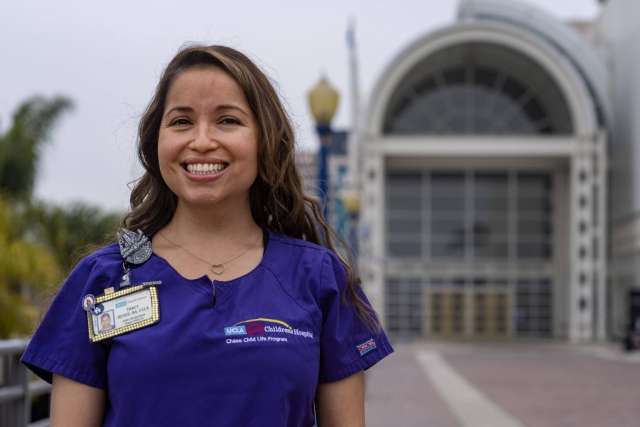 Person wearing purple scrubs with a hospital badge, standing outdoors in front of a large building with an arched glass entrance.