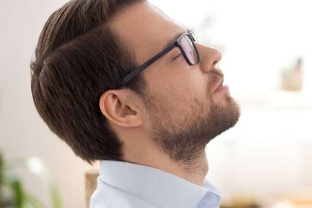 Side view of a person with short, neatly combed hair wearing a light-colored collared shirt. The background is softly lit with blurred indoor elements.