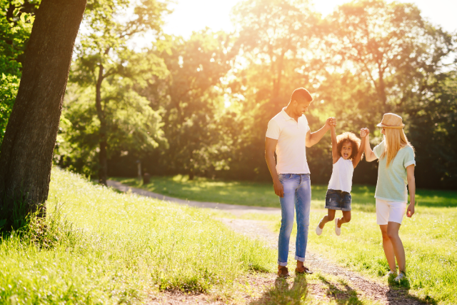 a father, child, and mother holding hands in an outdoor setting. there are grass, trees, and sunshine.