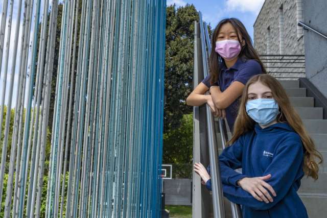 Two girls pose in a modern outdoor setting, one wearing a mask, near a blue installation.