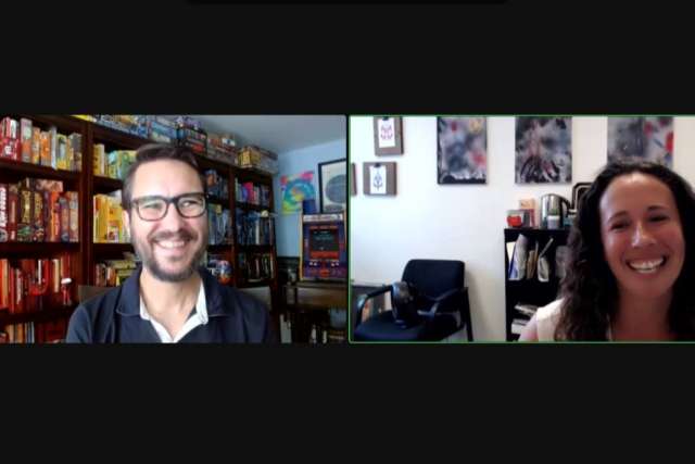 A screenshot of a video call showing a man with glasses and a shelf of board games in the left frame, and a smiling woman with abstract art in the right frame.