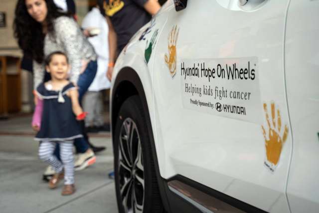 A toddler walks past the side of a white car with a "Hyundai Hope On Wheels" logo and orange handprints.