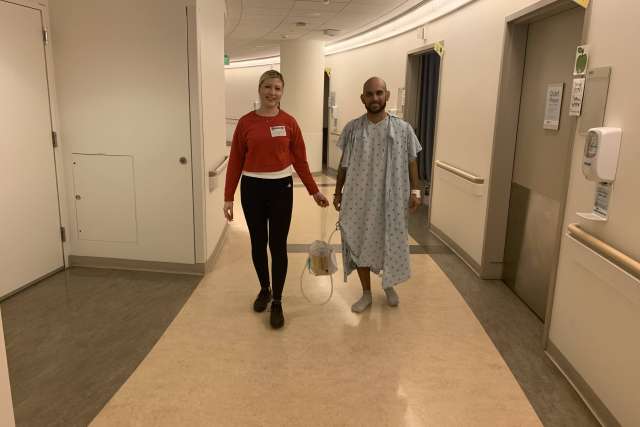 Two people walking down a hospital hallway, one wearing a patient gown and carrying medical equipment.