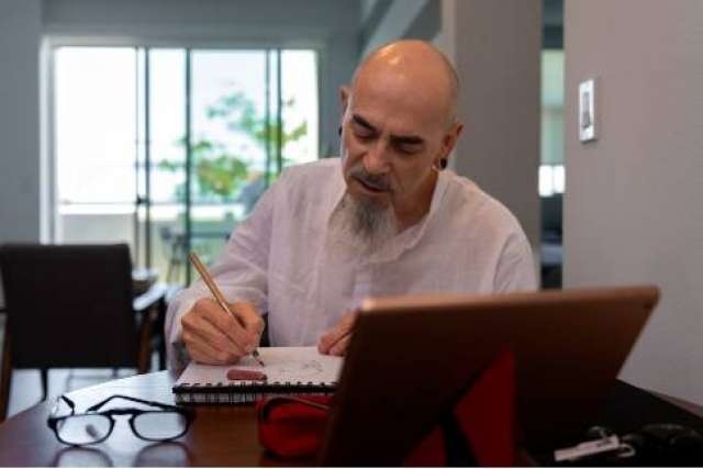 A focused headshot of Felix Baltazar, a bald man with a goatee, sitting at a table and writing in a notebook next to a tablet.