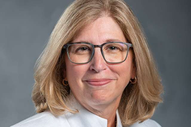 Headshot of a woman with blonde hair, wearing a white jacket and a decorative pin.