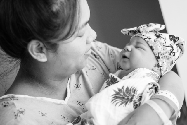 A black and white portrait of Ann Fortes holding her baby, Magnolia. The baby is swaddled in a blanket and wears a beanie with a bow.