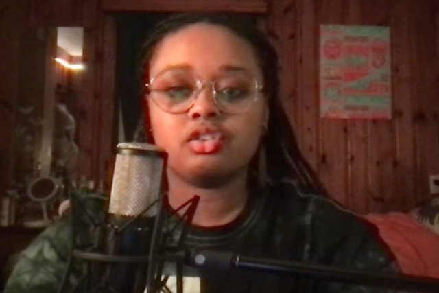  A young woman with braids and glasses speaks into a large studio microphone, with a wooden wall behind her.