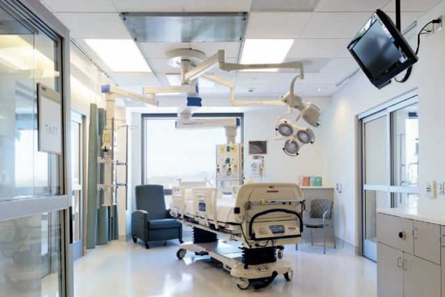an intensive care unit room that has a hospital bed, some lights and medical equipment hanging from the ceiling, a rest chair, and a monitor in one of the walls.