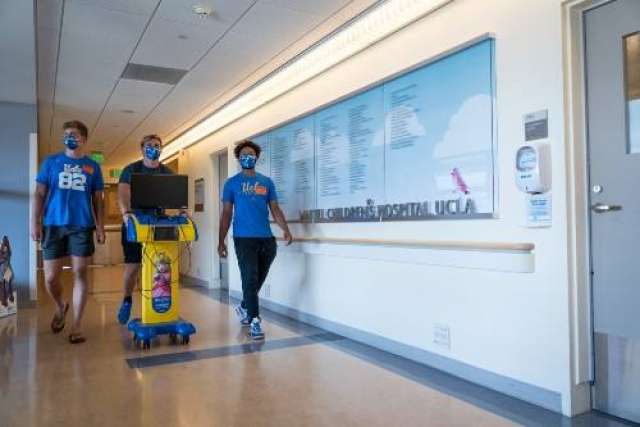 Masked staff in blue shirts push a yellow cart down a hallway