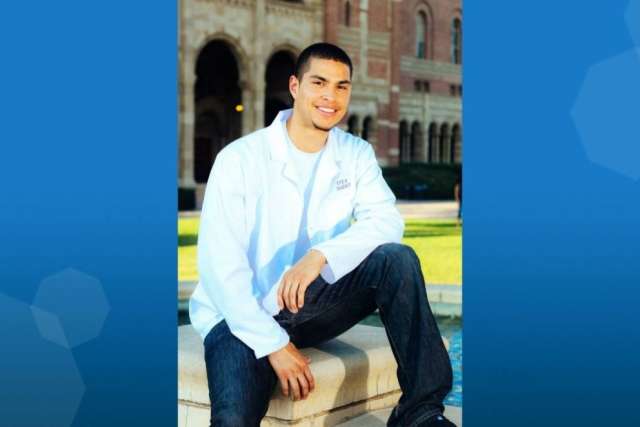 Person wearing a white lab coat and dark jeans seated on a stone ledge in front of a historic building with arches.