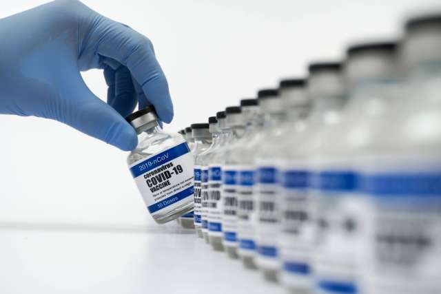 A gloved hand holds a vaccine vial in front of a row of similar vials.