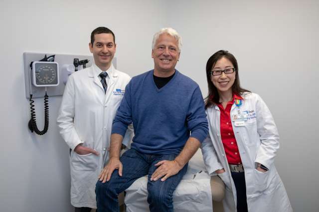 Two healthcare professionals in white coats standing beside a seated patient in a clinical setting with medical equipment on the wall.