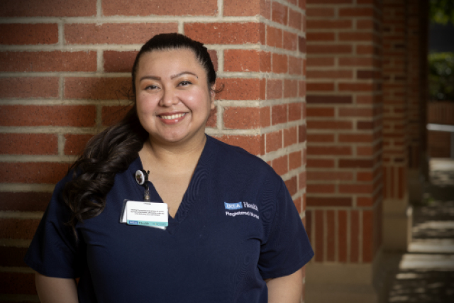 Dahlia Maldonado in navy blue scrubs stands in front of a brick wall, looking directly at the viewer and smiling.