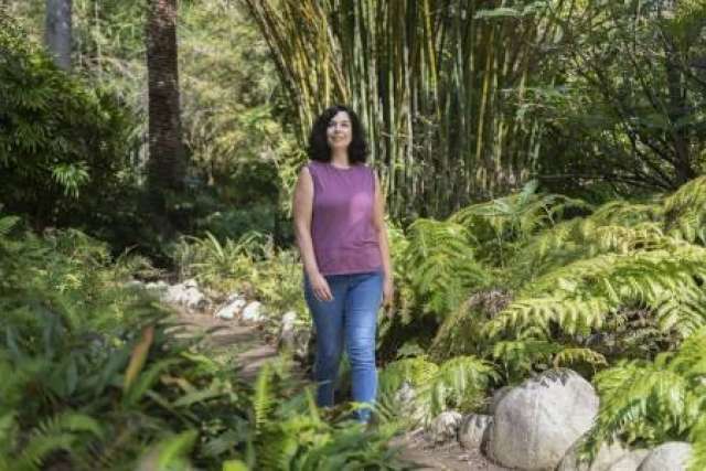 Caitlin Musick in a purple sleeveless shirt and jeans walks forward on a path in a lush, green forest.