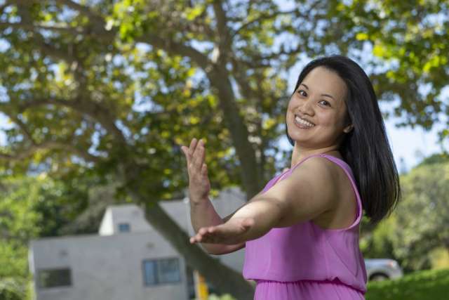 A smiling Janet Lo, a woman with dark hair, stands with her arms outstretched, as if dancing, on a sunny lawn.
