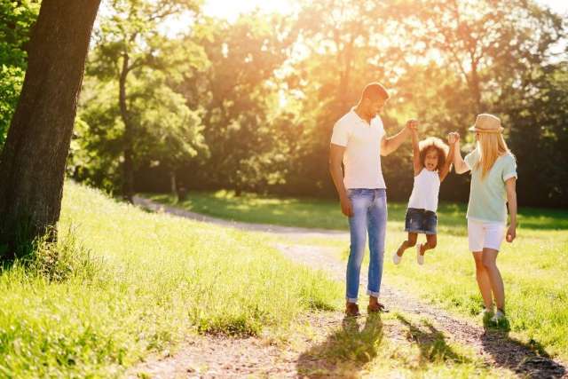 a father, child, and mother holding hands in an outdoor setting. there are grass, trees, and sunshine.