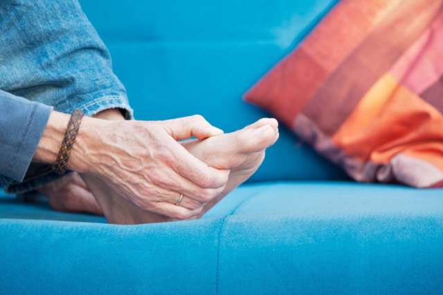 Person sitting on a blue couch holding their foot, appearing to check or massage toes near a colorful pillow.