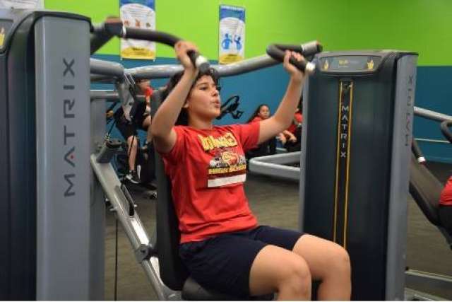 A female middle school student wearing a red t-shirt works out on a chest press machine.