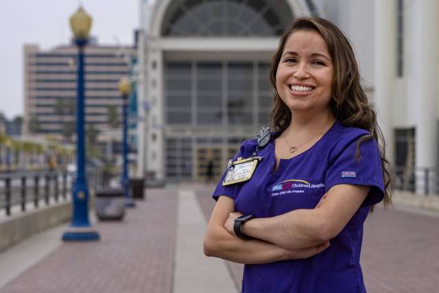 Person wearing purple scrubs with a hospital badge, standing outdoors in front of a large building with arched glass entrance.
