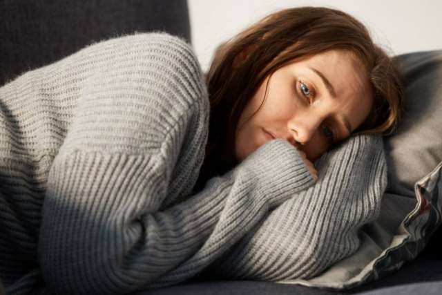 Person lying on a couch curled up in a gray knit sweater, resting on a pillow with natural light coming from the side.