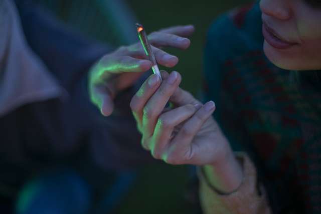Person holding a cannabis cigarette, with another hand reaching towards it in dim lighting.
