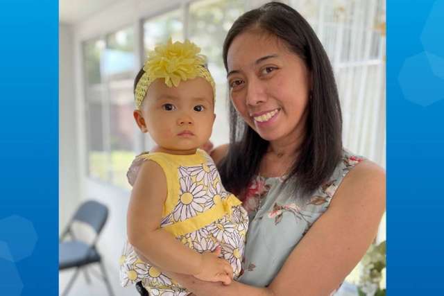 A woman holds a child outdoors, both smiling, with bright natural lighting.
