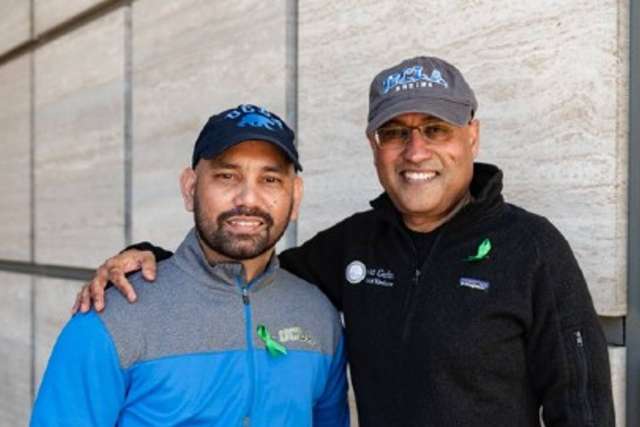 Two people wearing UCLA-branded caps and jackets with green awareness ribbons, standing side by side against a light stone wall