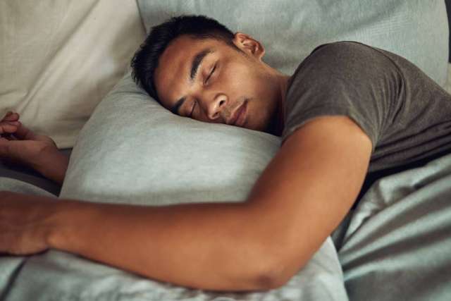 Person lying face down on a bed, wearing a dark short-sleeve shirt, with arms extended and resting on light-colored pillows and bedding.