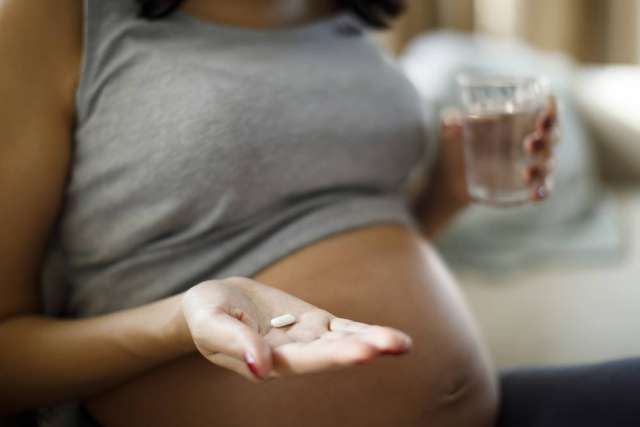 A close-up of a pregnant woman in a gray tank top holding a glass of water in one hand and two pills in the palm of the other, above her baby bump.