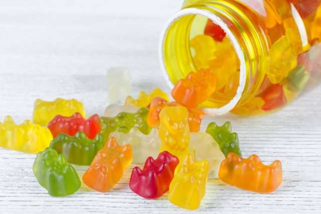 Colorful gummy vitamins or candies shaped like bears are spilled out of a yellow plastic bottle onto a white wooden surface.