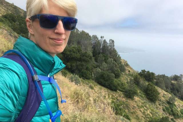 Lori Jordan, a white woman with short blonde hair and sunglasses, smiles while hiking. She wears a teal puffer jacket and a backpack.