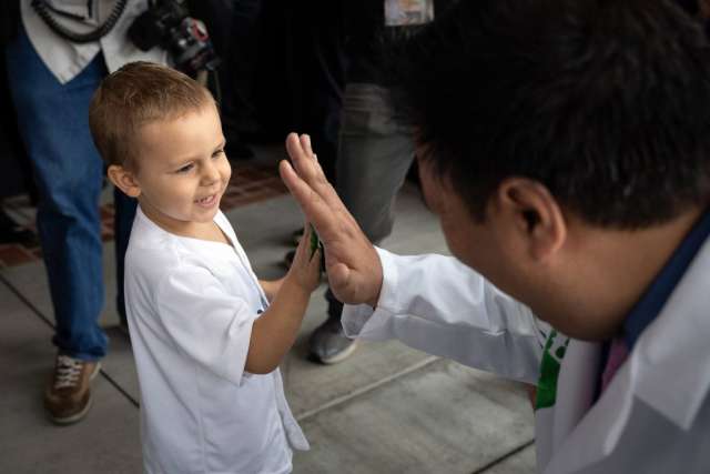 A young boy in a white shirt smiles as he high-fives an adult in a white lab coat, seen from behind.