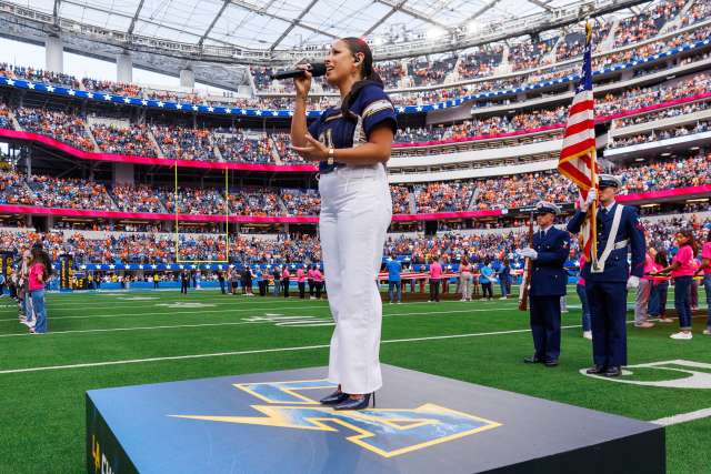 A woman with a microphone stands on the podium on the field in front of a larger crowd before a Los Angeles Chargers game.