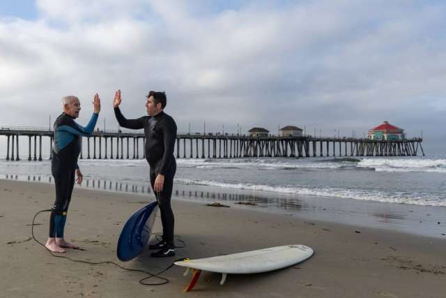 Dr. Sassoon and Robert Lombard high fiving on the beach beside their surfboards