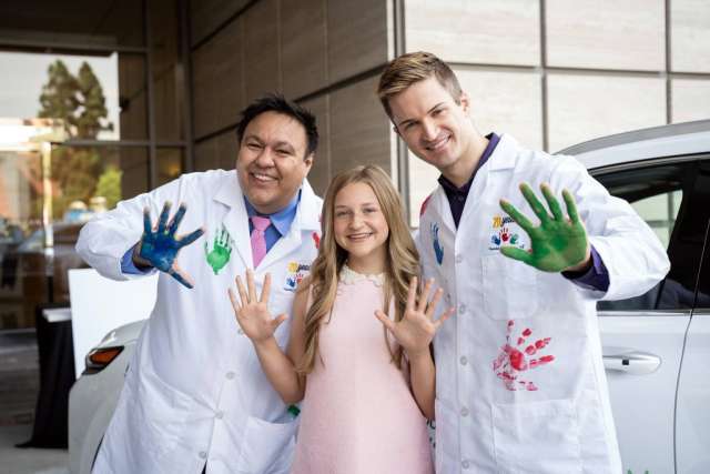  Two men in lab coats, one with blue-painted hands and one with green, pose with a smiling girl in a pink dress, all showing their painted hands.
