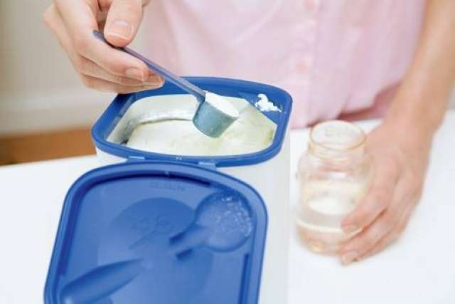 A person scoops infant formula from a container with a measuring spoon.