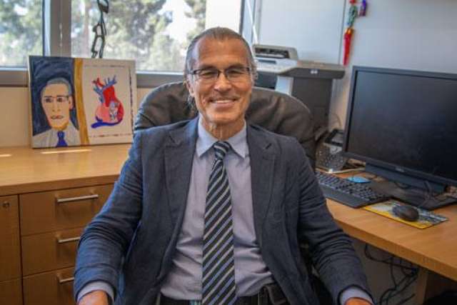 Man seated in an office, wearing a suit with artwork on the desk behind him.