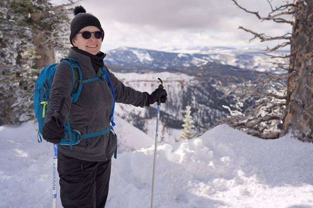 Laurie Adami winter hiking gear, wearing a hat, sunglasses, and a backpack, smiles while holding ski poles in a snowy mountain landscape.