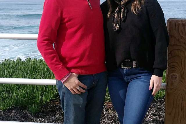 Two people standing outdoors near a white railing with ocean waves and greenery in the background, wearing casual sweaters and jeans.