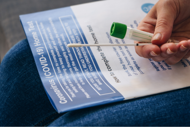 A person holding a cotton swab and COVID test tube in their lap, alongside a flier with COVID at-home test directions
