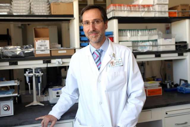 A middle-aged man in a white lab coat, glasses, and a colorful striped tie stands in a busy laboratory setting, smiling at the camera.