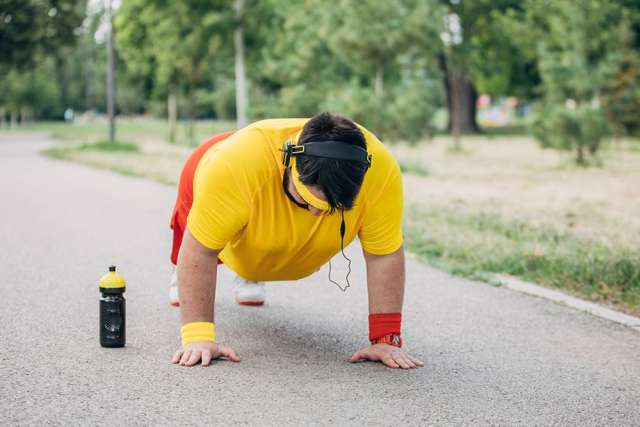 A person wearing a yellow shirt and red shorts is doing a push-up on a paved path in a park, with a water bottle placed nearby and headphones on.