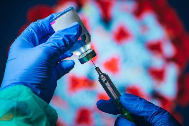 Gloved hands drawing a COVID-19 vaccine dose from a vial with a syringe, with a virus illustration in the background.