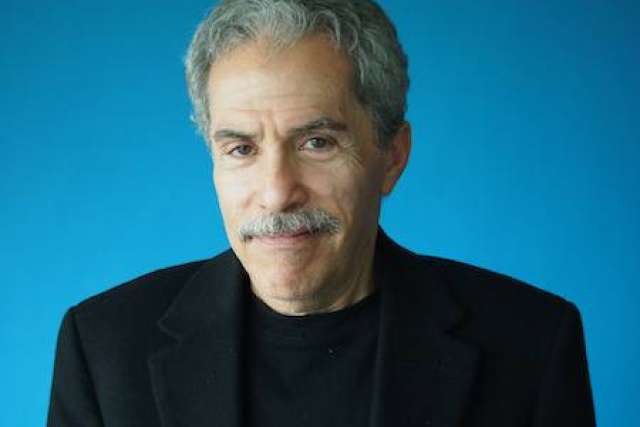 Portrait of Michael Strober, PhD with gray hair and mustache wearing a black jacket and shirt against a blue background.