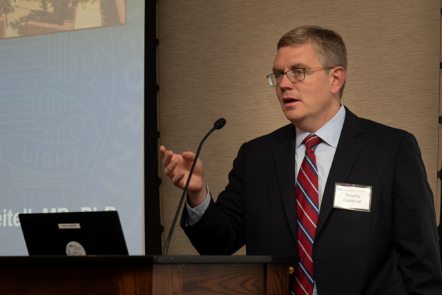 Dr. Donahue, in a suit and tie, wearing glasses, stands at a podium with a microphone, gesturing with his hand while speaking.