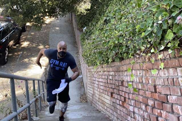 A muscular man in a dark t-shirt with text runs up a concrete staircase next to a brick wall with green ivy. A black SUV is at the top.