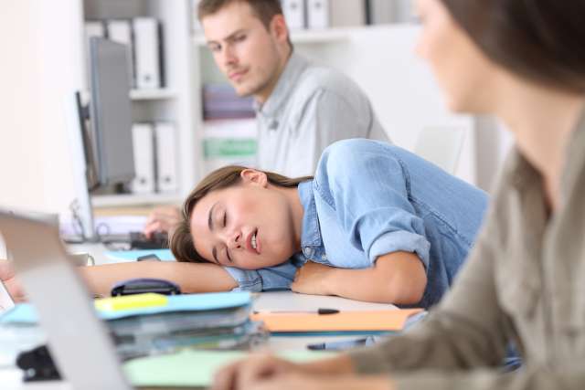 Woman sleeping with head on desk in classroom while coworkers look at her with concerned expressions.