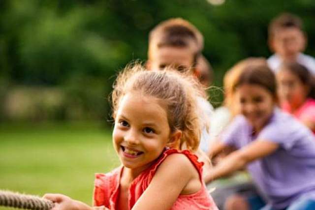 Children playing tug-of-war outdoors, pulling on a thick rope in a grassy area with trees in the background.